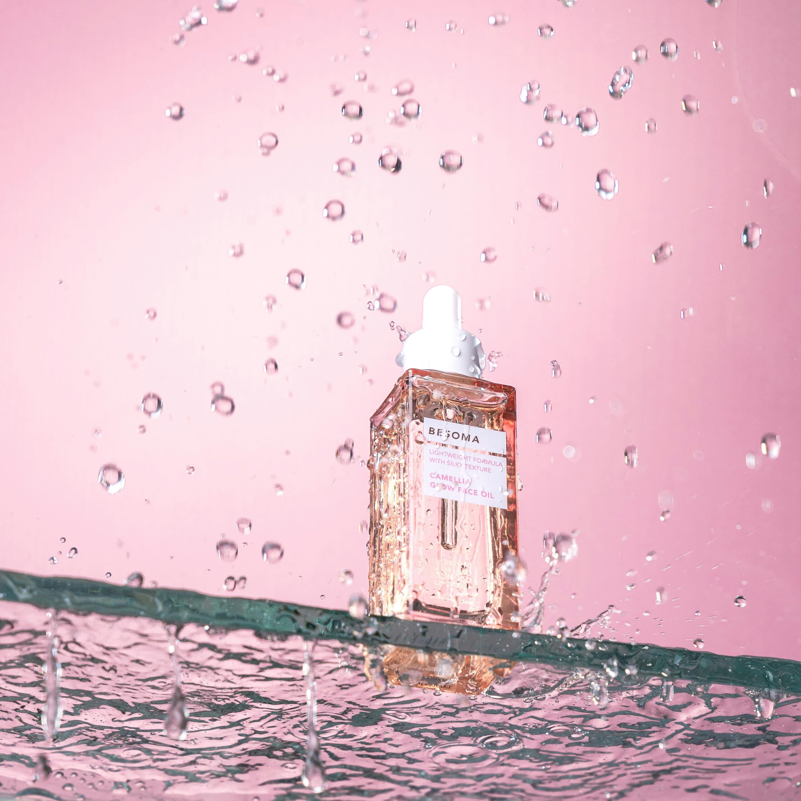 A studio shot of an elegant cosmetic serum with water splashes on a pink background.