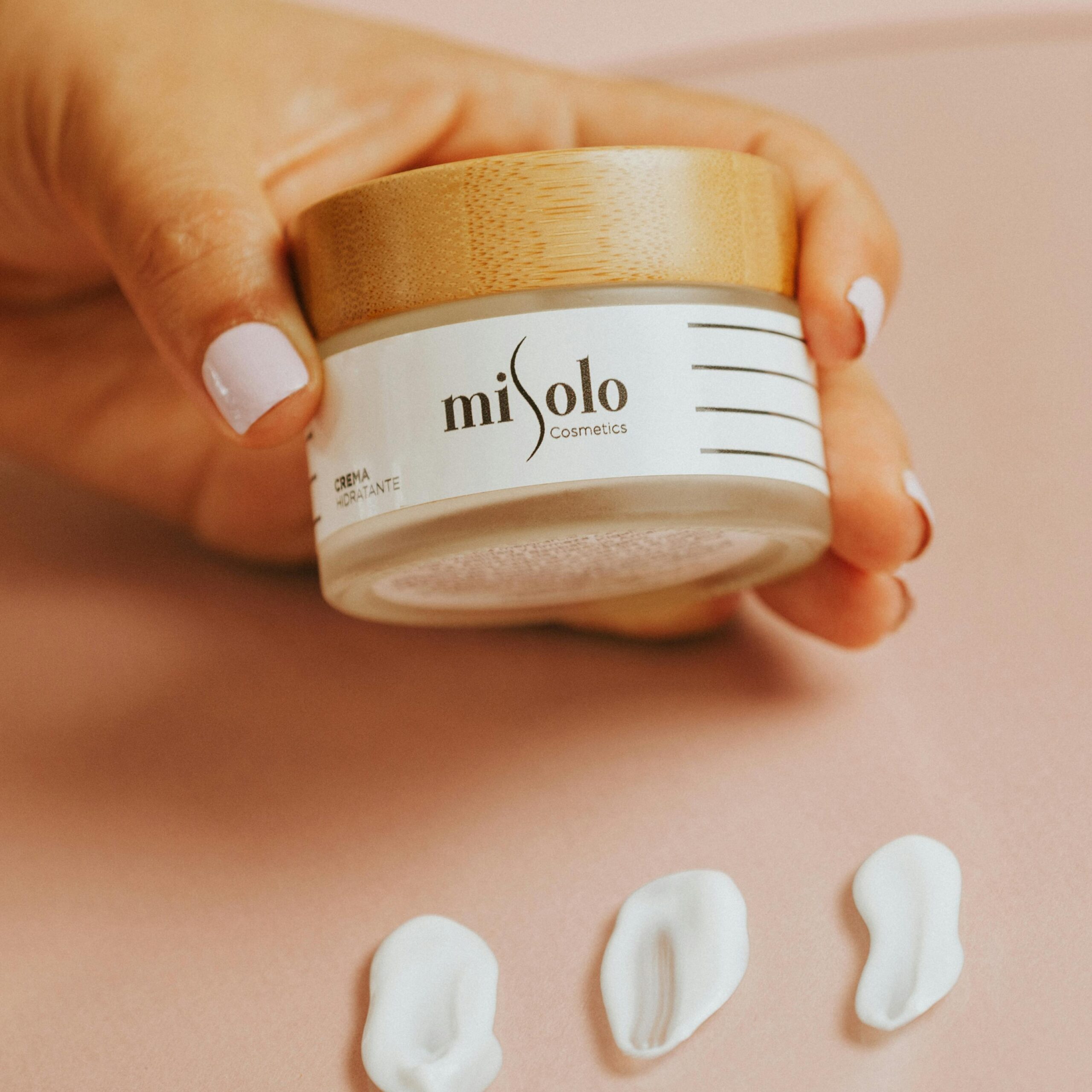 A hand with manicured nails holding Misolo cosmetic cream in a stylish jar on a soft pink background.