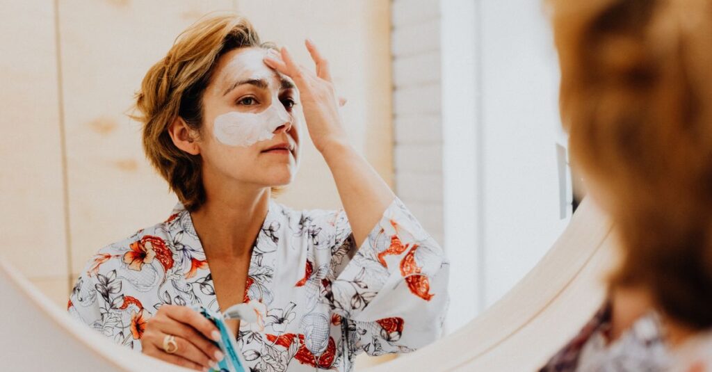 Adult woman applies facial mask while looking in bathroom mirror, promoting skincare routine.