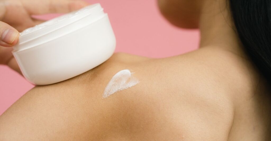 Skincare Woman applying skincare cream to shoulder against a pink background.
